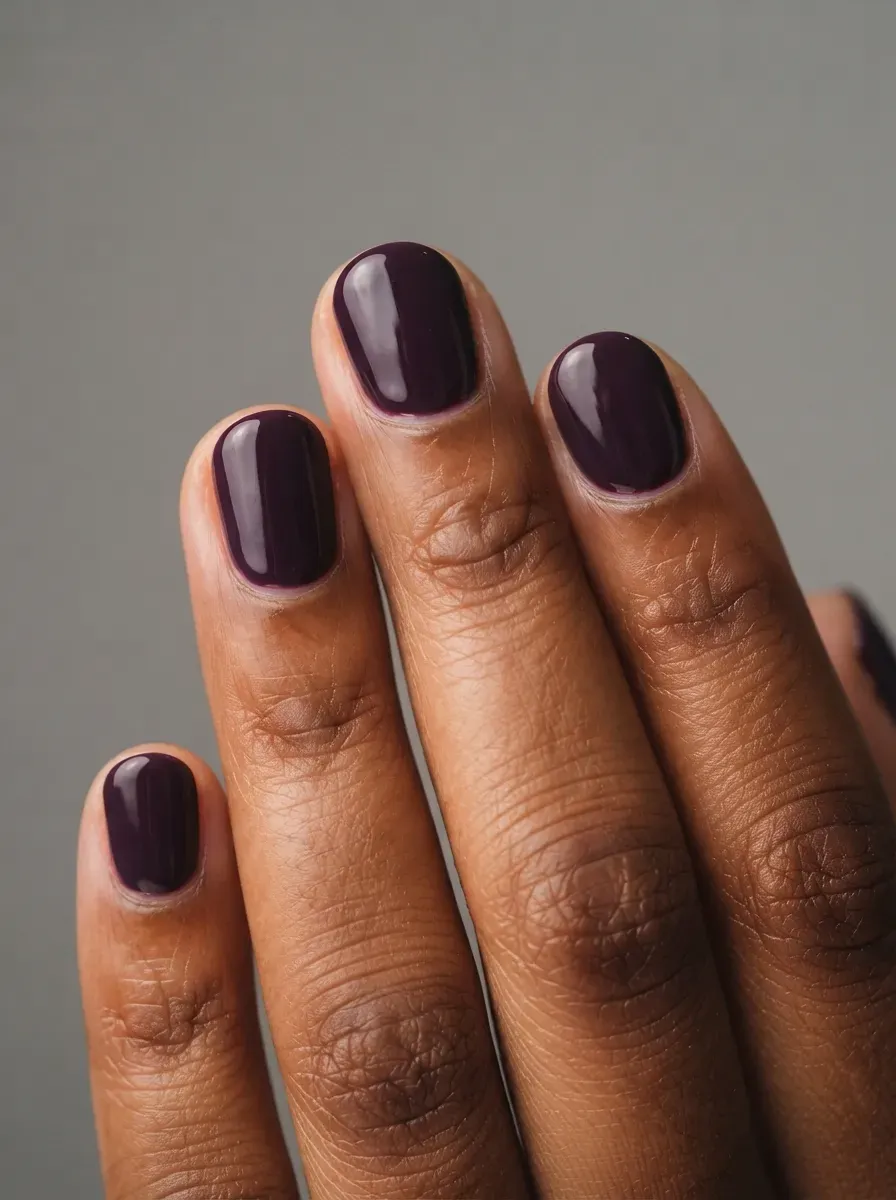 Short rounded nails with glossy deep plum polish on brown skin