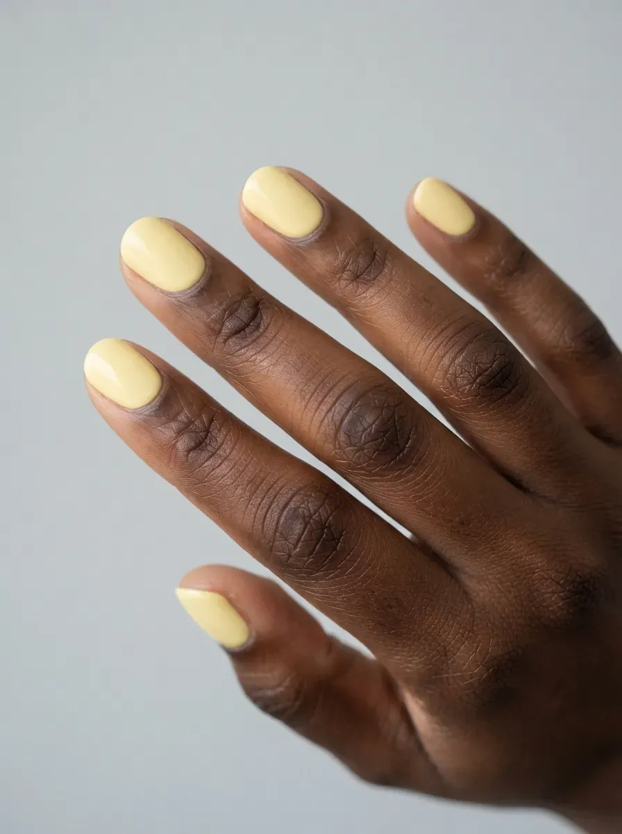 Close-up of short round nails in pale butter yellow creme on rich dark skin tone